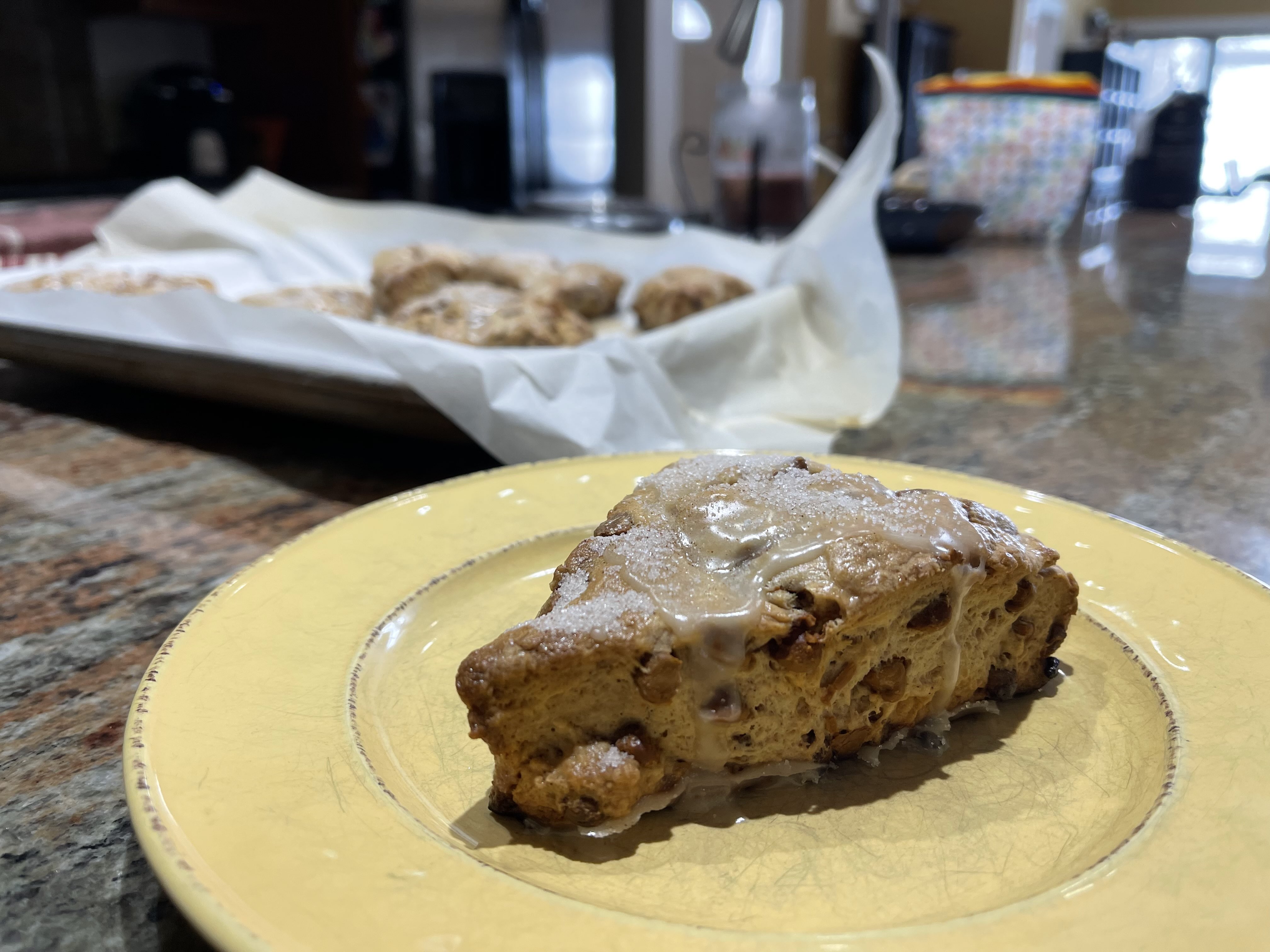 A cinnamon crunch scone on a plate with a baking sheet of them behind.