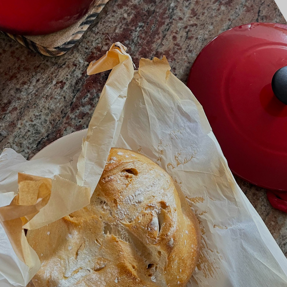 A rustic sourdough loaf on some parchment with a dutch oven lid to the right